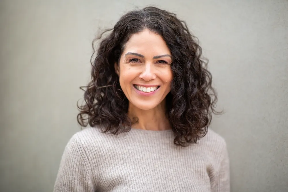 A woman with curly hair smiling, against a pale grey wall
