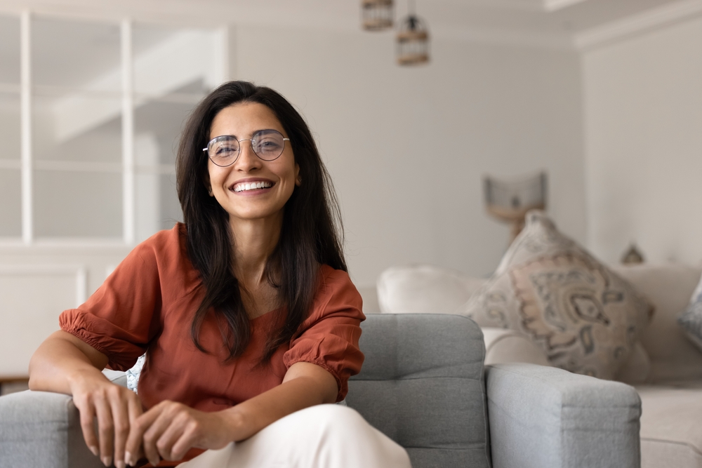 A woman in glasses and a rust-coloured top, sitting on a sofa