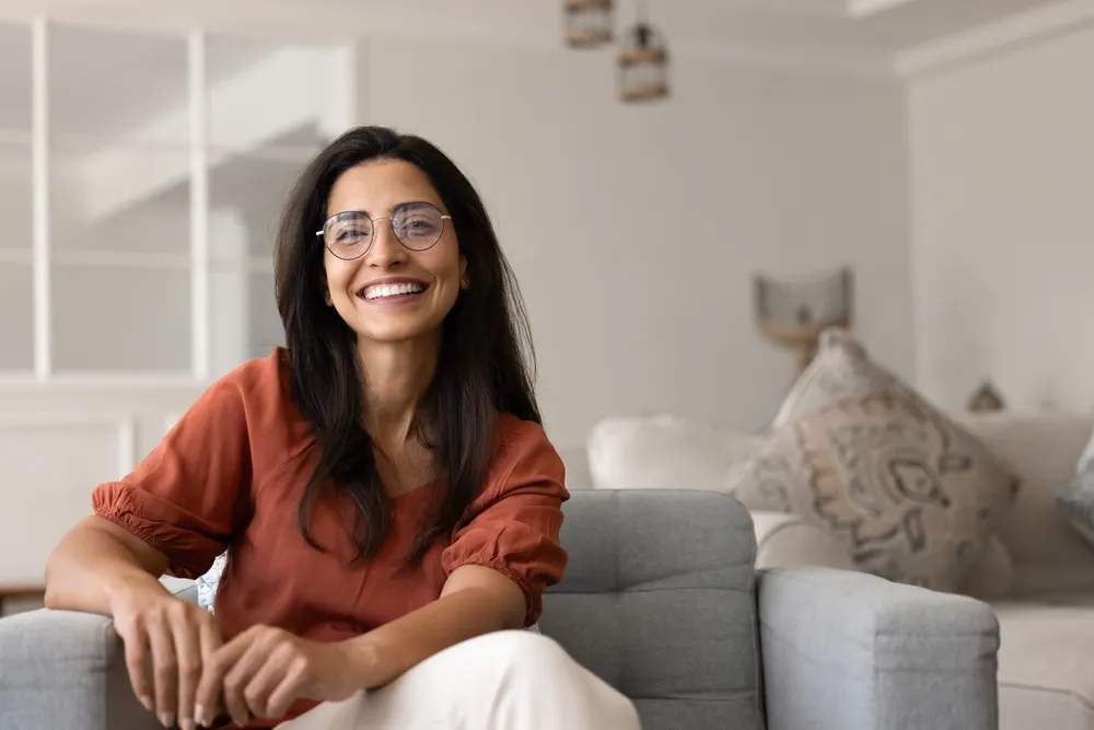 A woman in glasses and a rust-coloured top, sitting on a sofa