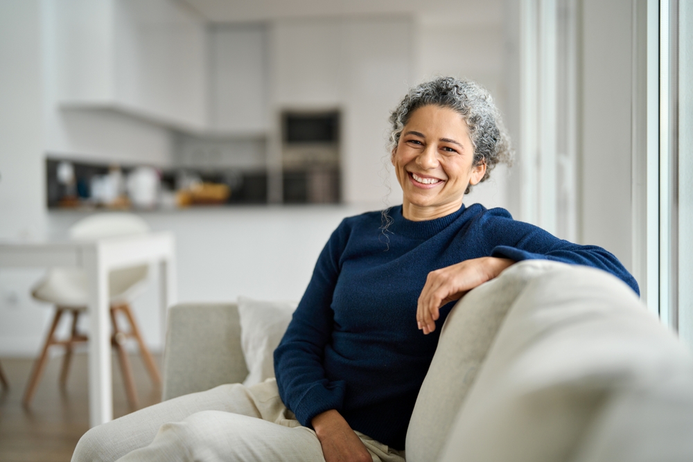 A woman with grey curls in a navy jumper, sitting on a sofa and smiling