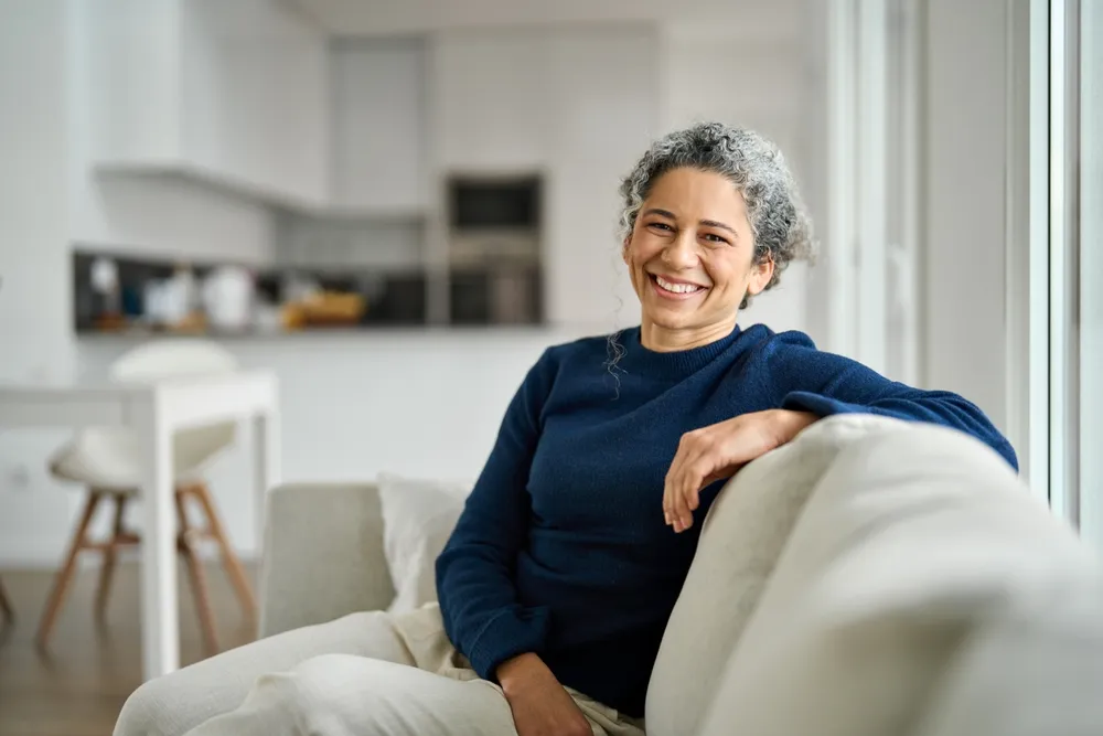 A woman with grey curls in a navy jumper, sitting on a sofa and smiling