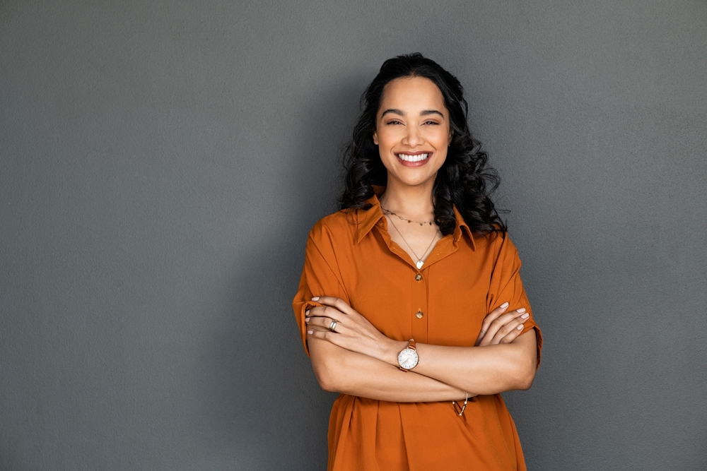 A woman smiling with her arms folded, against a grey wall