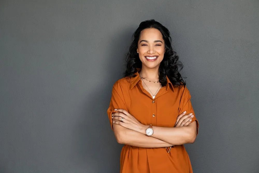 A woman smiling with her arms folded, against a grey wall